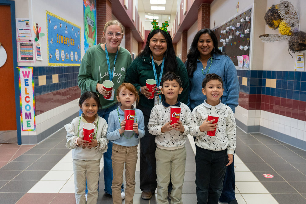 three women and four young kids standing in school hallway. The kids are hold Raising Cane's cups.