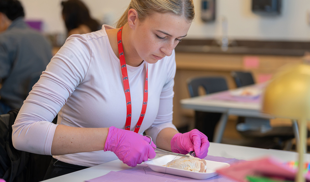 teen girl with long blond hair wearing pink gloves works on sutures on a piece of chicken in classroom