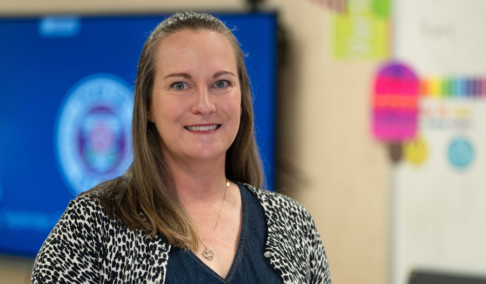 woman with long brown hair and printed jacket standing in classroom