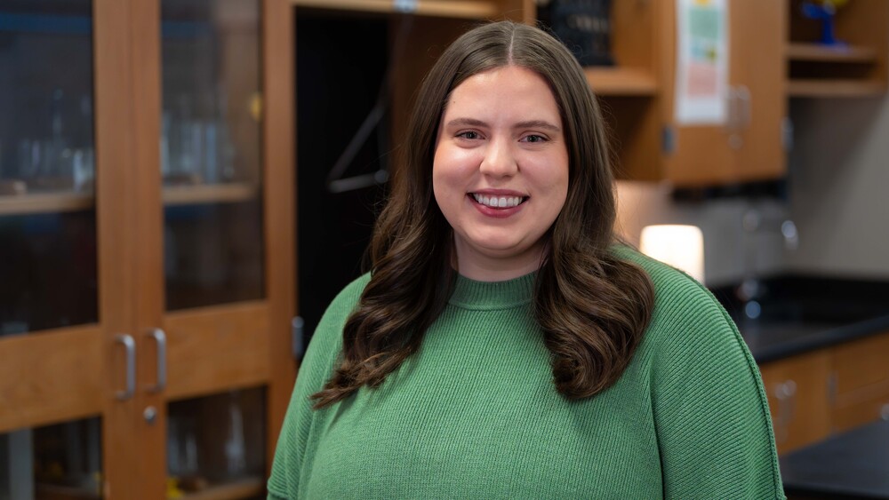 woman with long, dark hair wearing green sweater standing in front of cabinets in classroom