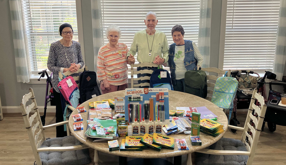 elderly man and and three  women stand behind table with school supplies on it