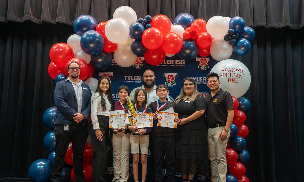kids holding certificates, trophies, and medals standing next to men and women with a balloon arch around them