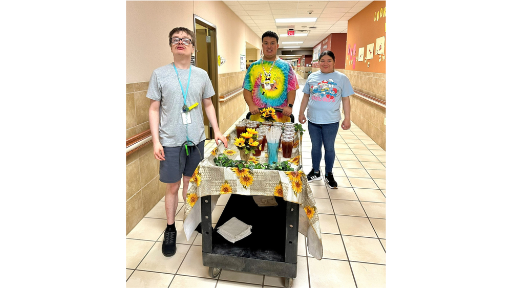 two teen boys and a teen girl pushing a cart with tea down a school hallway