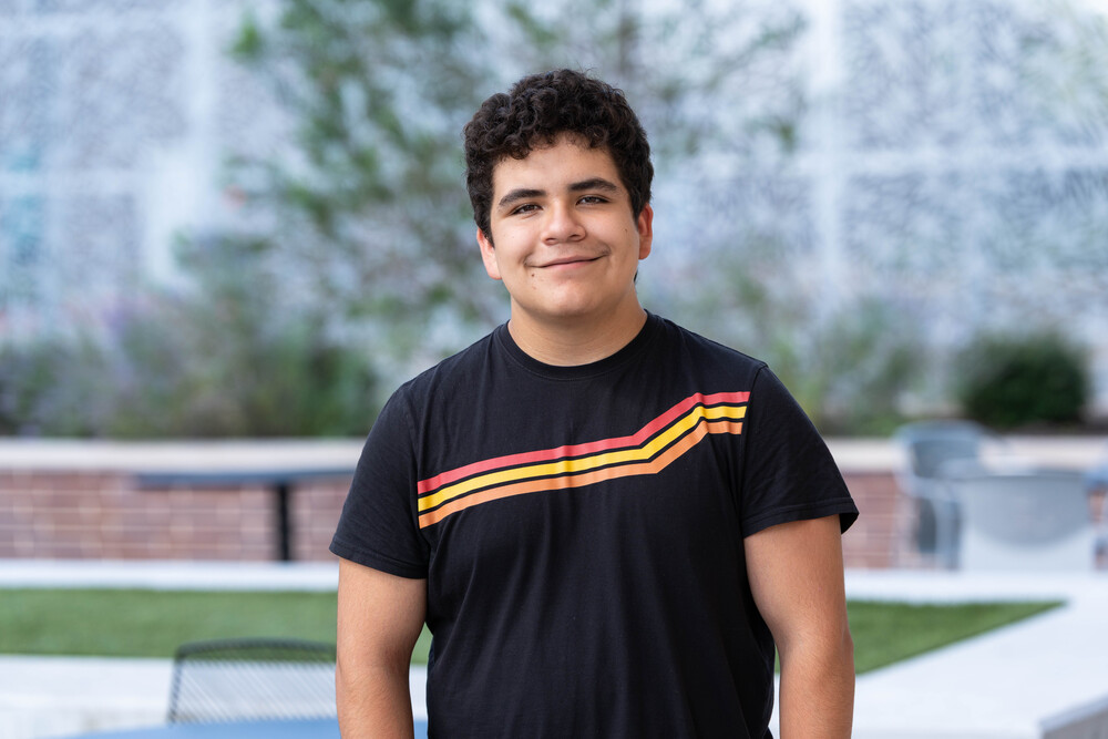 Hispanic teen boy wearing t-shirt with stripes across chest standing in courtyard at school