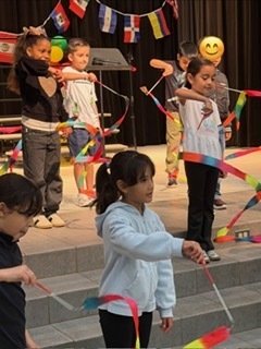 Seven elementary students practice on stage for the Hispanic Heritage Program, twirling colorful ribbons as international flags hang in the background