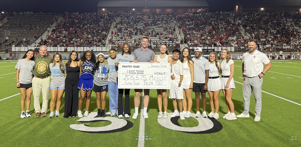 men, women, and teens standing on 50-yard line at football stadium holding a giant check