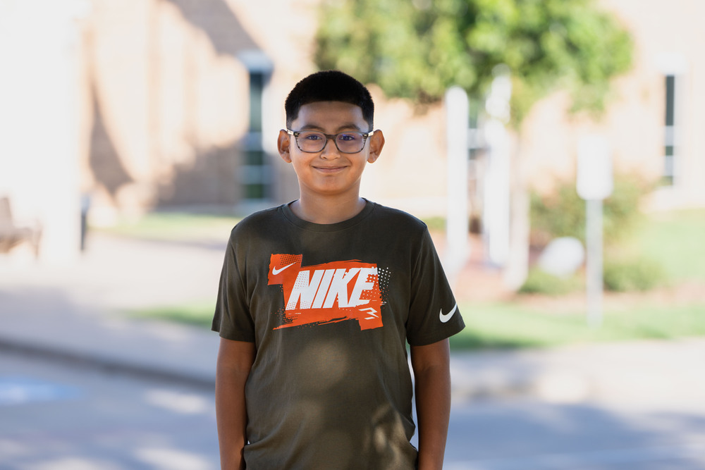 elementary age boy wearing glasses and a gray t-shirt standing outside a school