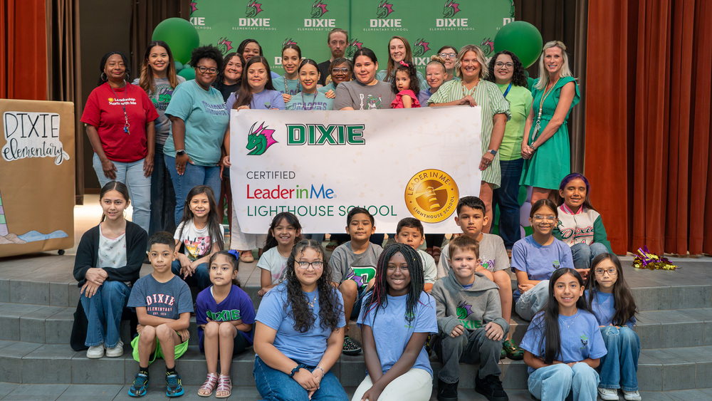 adults and children on stage holding a Dixie Certified Leader in Me Lighthouse School banner