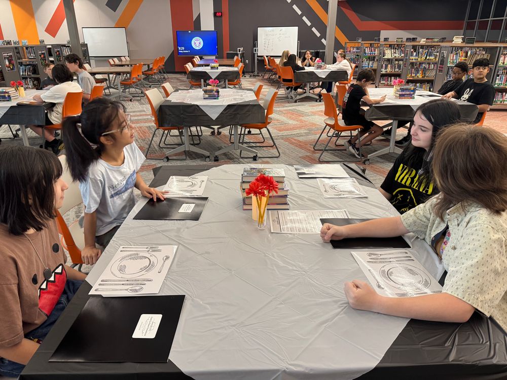 multiple groups of students sitting at a tables decorated for a book tasting.