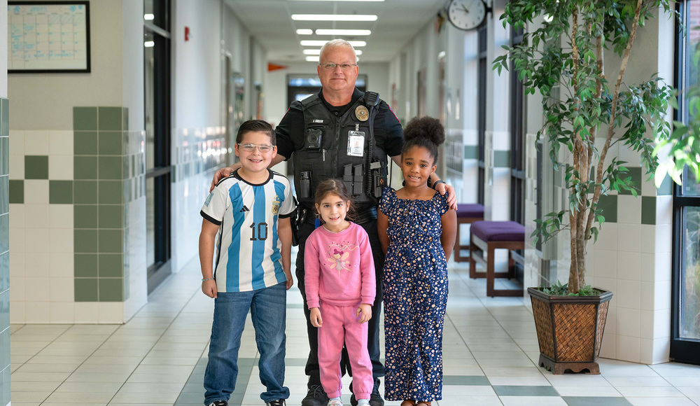 man in police uniform standing behind three elementary age kids in a school hallway