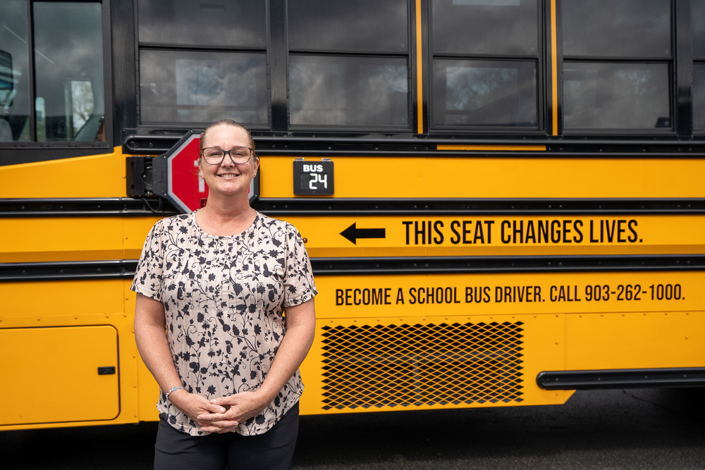 woman standing beside school bus. Side of bus says, "This seat changes lives. Become a school bus driver. Call 903-262-1000."