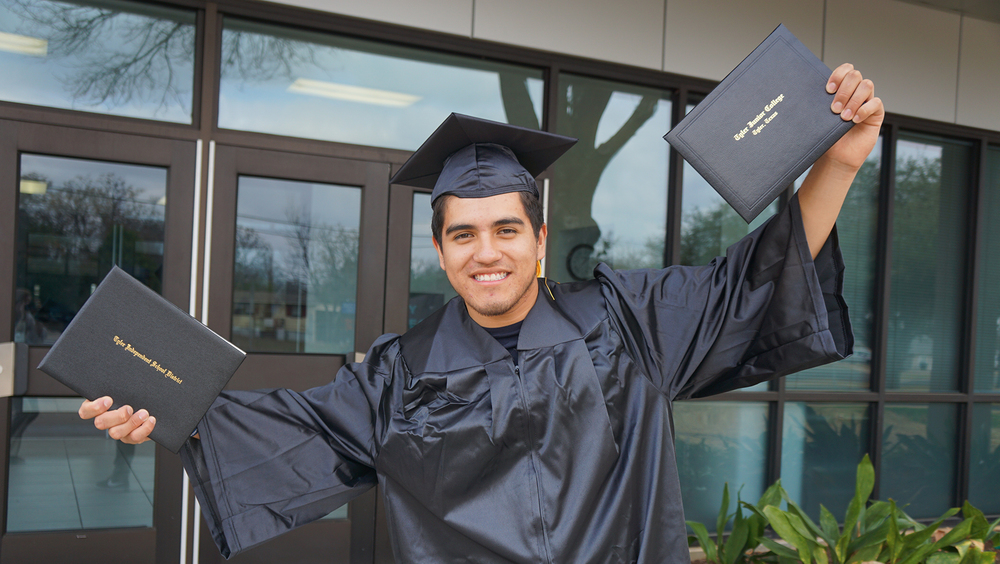 teen male with black graduation cap and gown on holding two diplomas