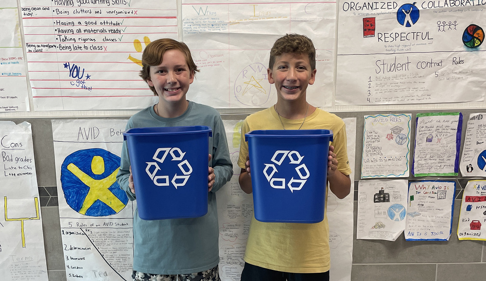 two teen boys standing in hallway holding blue recycle bins