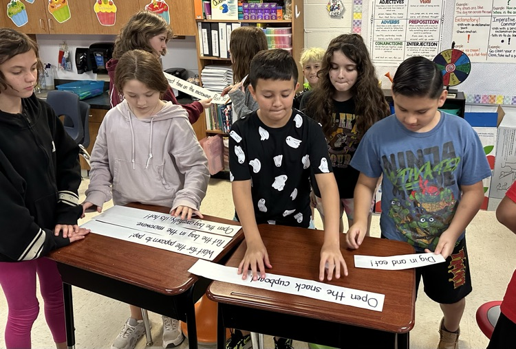 four students standing at a group of desks organizing sentence strips