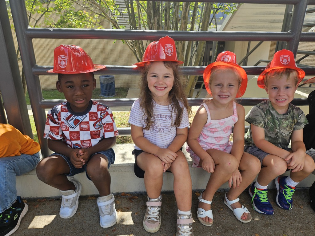 Students sitting on curb while wearing firemen hats.
