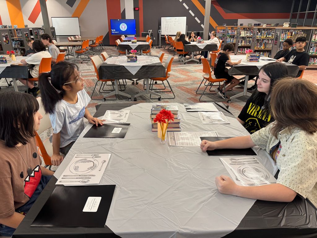 multiple groups of students sitting at a tables decorated for a book tasting.