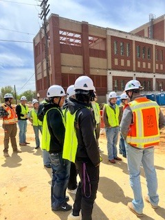 Students in safety gear listen to the Construction Site Superintendent discussing the work on an active job site. Large Building in the background. 