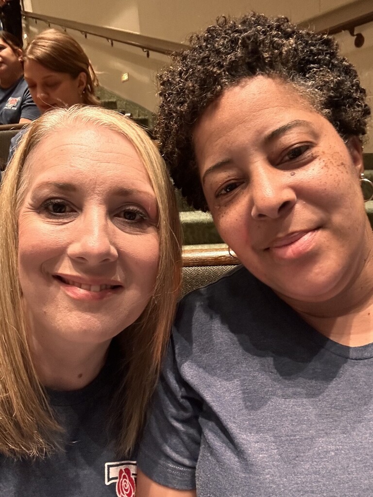 Two women in matching shirts, one with blonde hair and the other with curly hair, smiling.