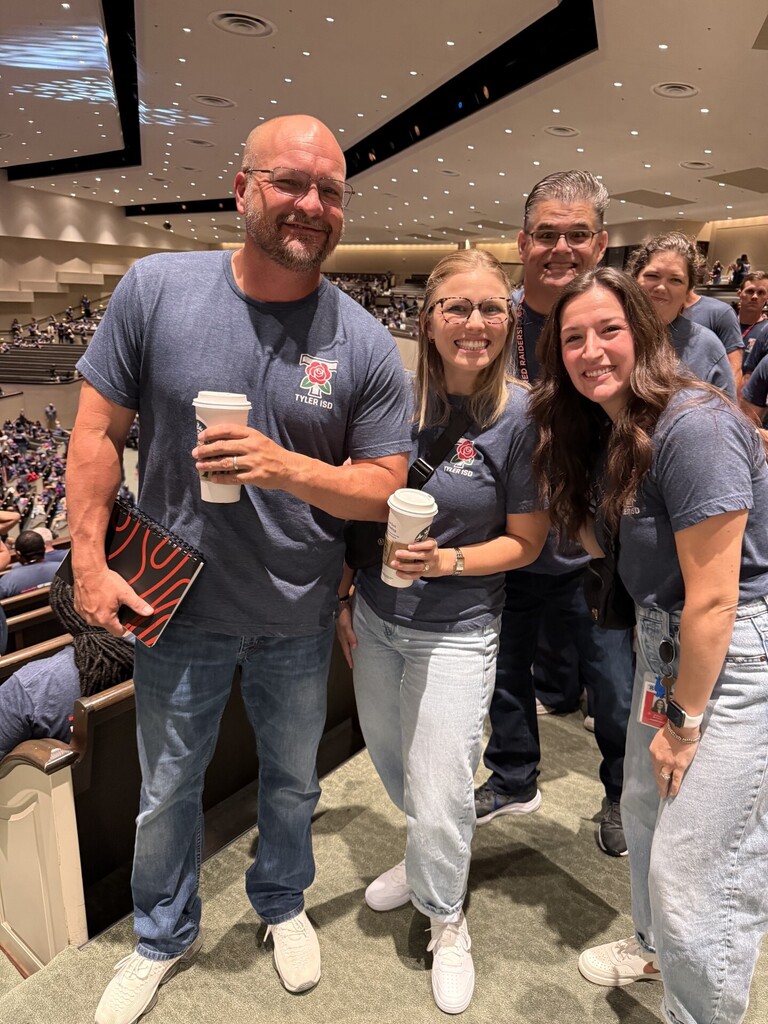 Group of people, smiling, in a conference hall. Two holding coffee cups, one with a book.