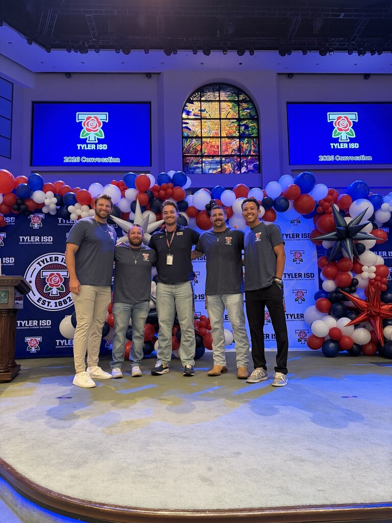 Five men stand on a stage, smiling. Behind them, large balloons form a decorative backdrop. Two large screens display text.