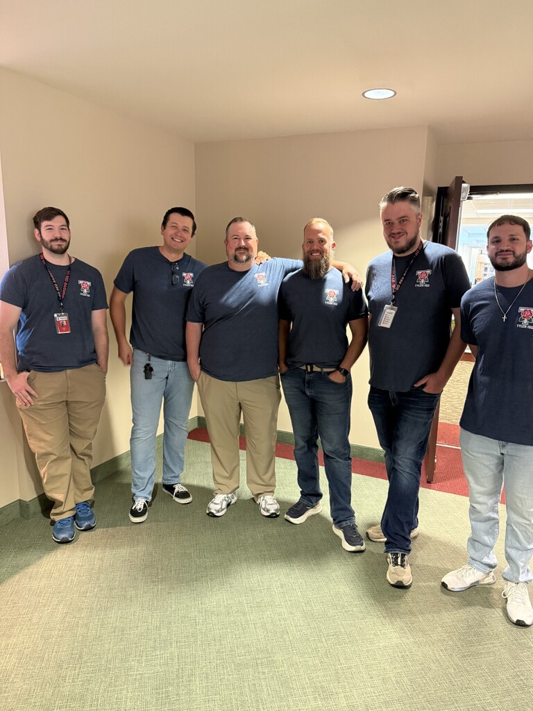 A group of men in matching blue T-shirts and jeans standing in a hallway with green flooring.