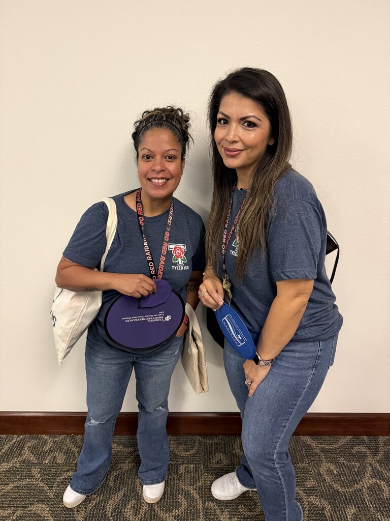 Two women in matching shirts and jeans pose for a photo. One holds a purple item.