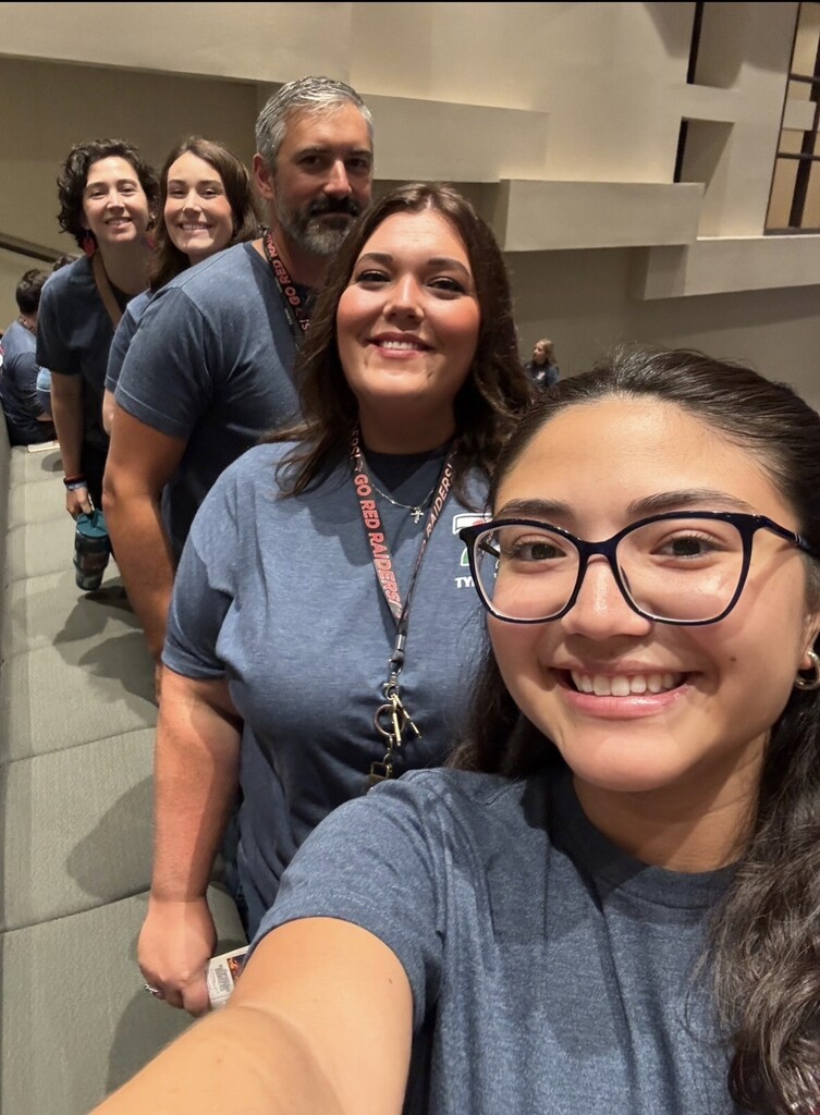 A group of people in matching blue shirts stand on stairs. A woman with glasses is taking a selfie.