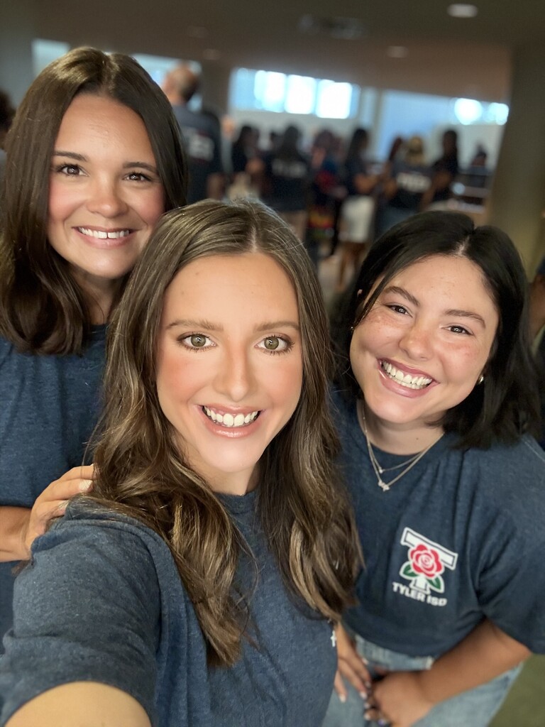 Three women smile for the camera, standing close together. They wear navy blue t-shirts. A crowd of people is blurred in the background.