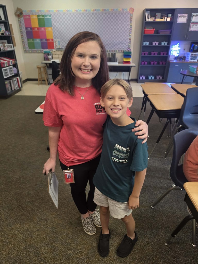 Woman and child standing in classroom. The woman wears a pink shirt, black pants, and black shoes. The child wears a green shirt, white shorts, and black shoes.