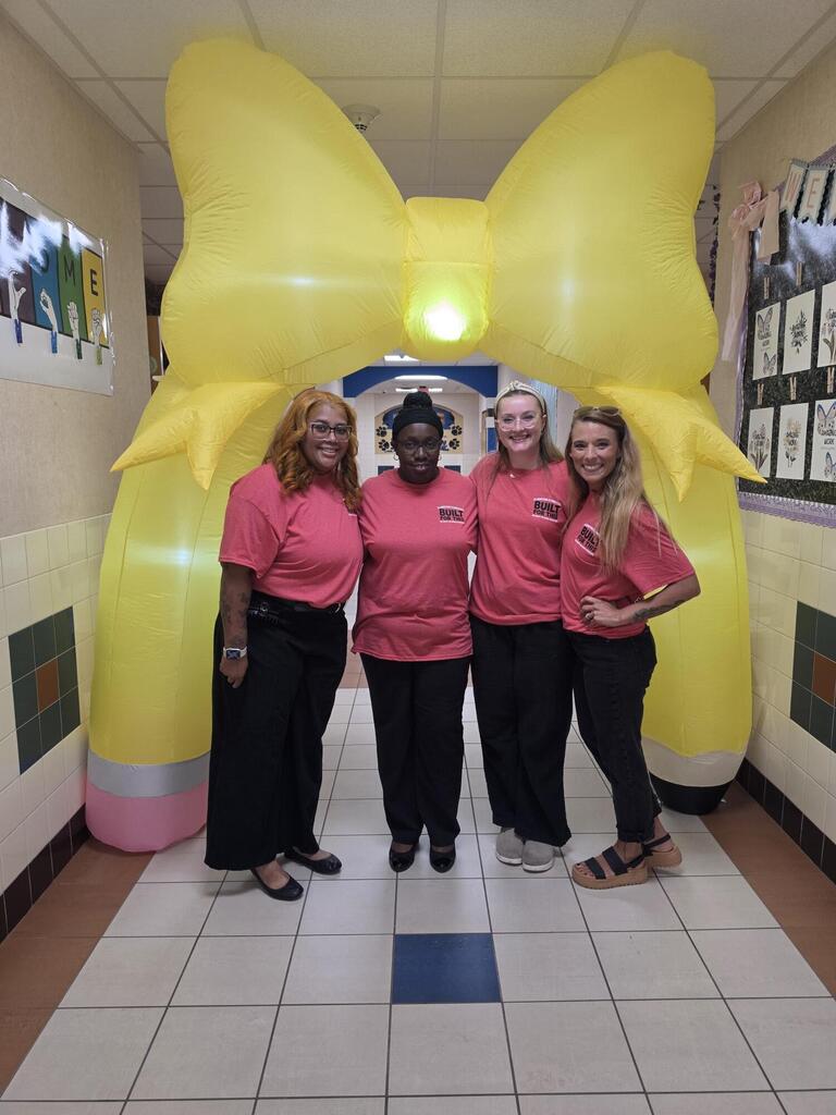 Four women in pink shirts and black pants pose before a yellow balloon bow. Behind them, a hallway with white tiles and framed pictures.