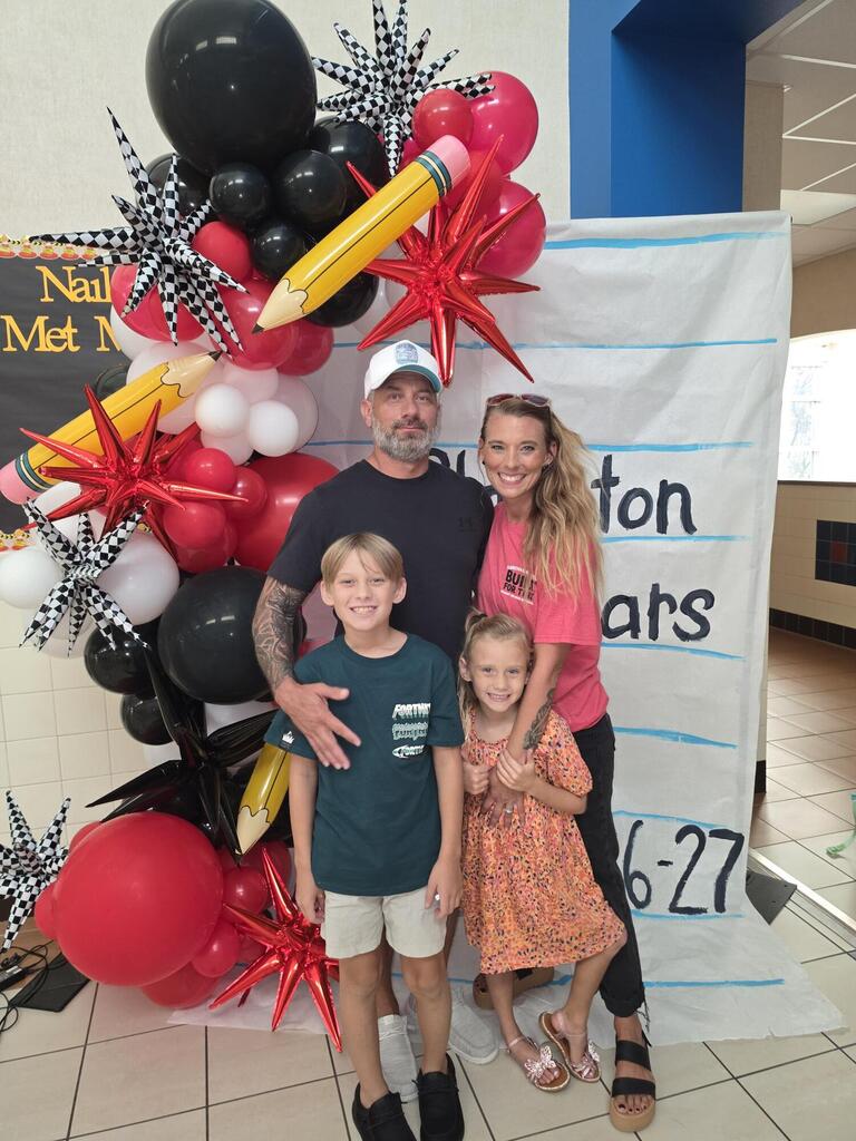 Four people stand together in front of a balloon backdrop with a "Newton Years 6-27" sign.