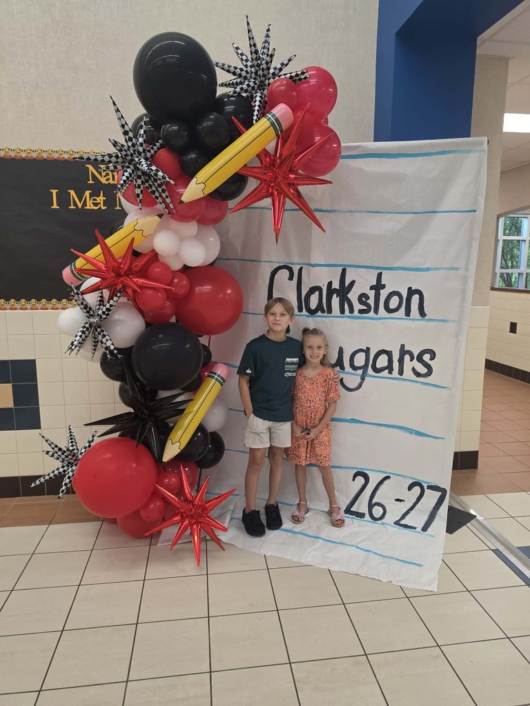 Two children stand near a large, decorative balloon arch with school-themed words and numbers.