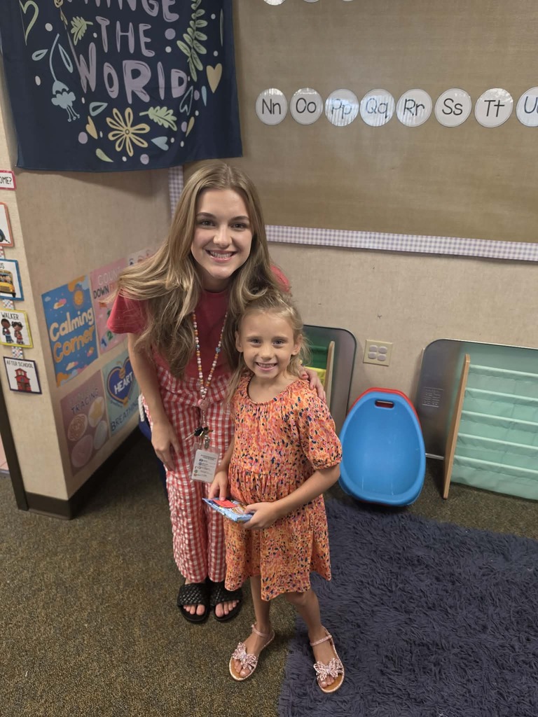 A woman and a girl stand in a classroom with a banner, colorful wall decorations, and a rug.