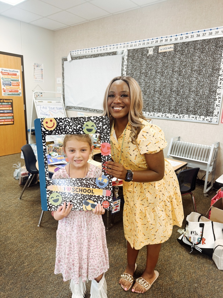 A woman in a yellow dress and a girl in pink dress stand in a classroom. The girl holds a photo frame with a smiley face.