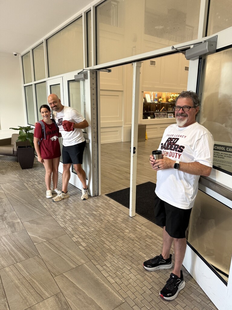 Three individuals pose by an open door. A man holds a cup. Behind them is a plant and a glass wall.