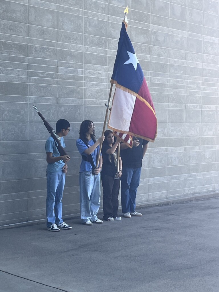 Four individuals standing in front of a textured wall, one holding a flag.
