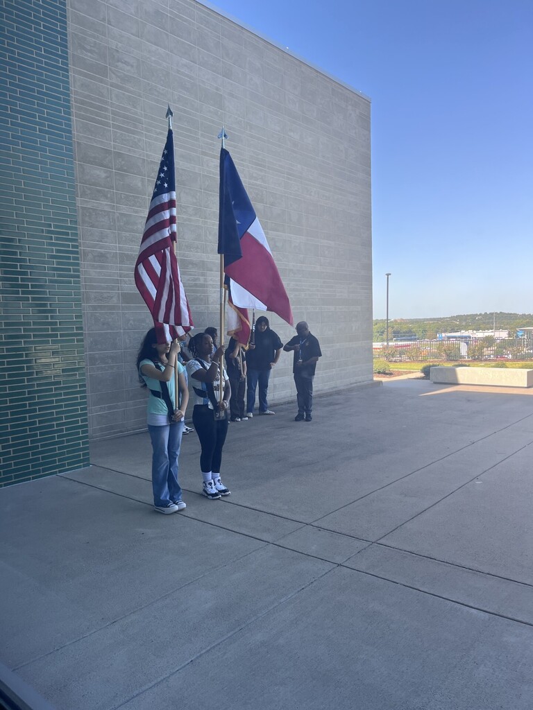 Several people stand outside near a building with two flags, one US flag and one Texas flag.