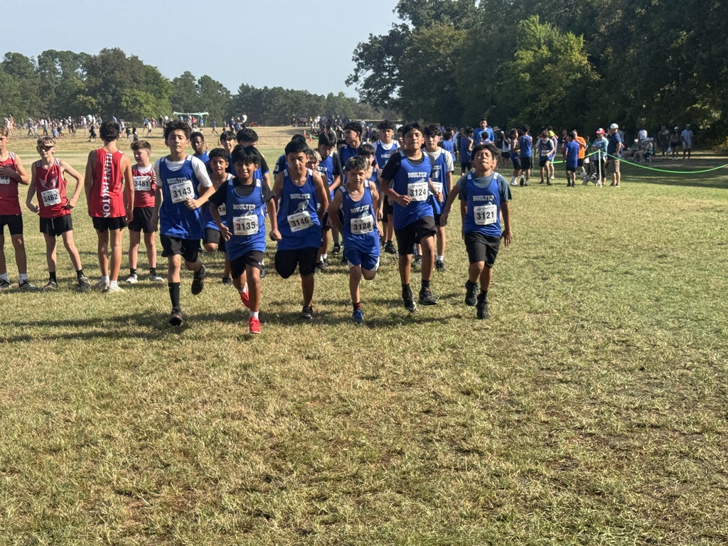 Group of people in blue shirts and numbers running in a grassy field with trees in background.