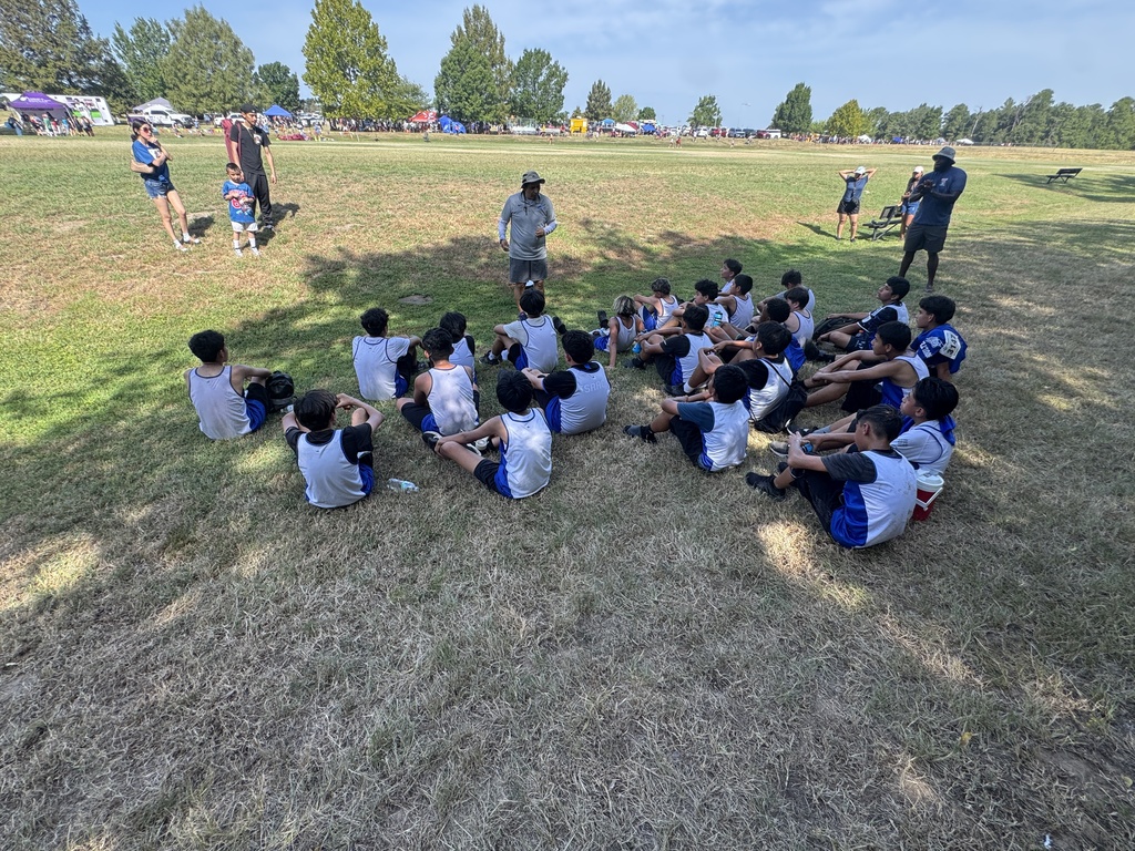A group of children wearing blue and white uniforms listens to a man in a field.