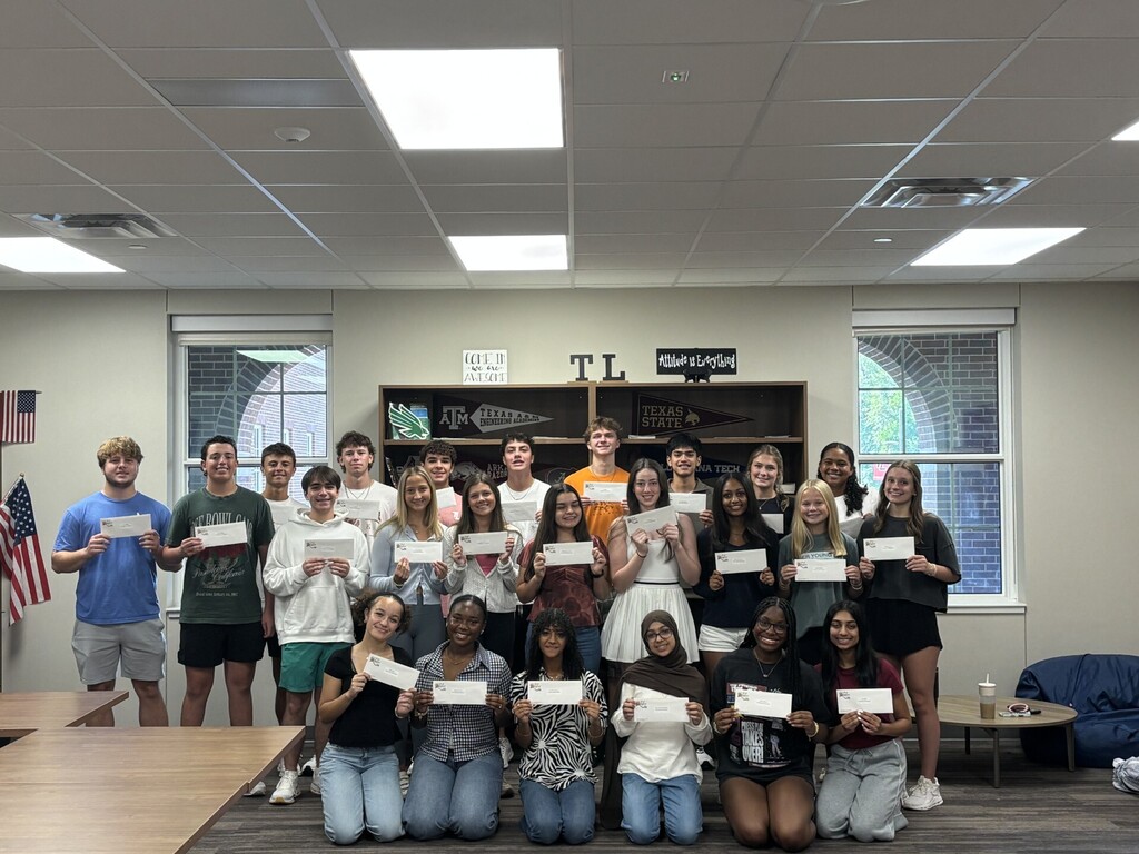 A group of students in a classroom holds certificates, standing and sitting. A flag is on the left, and windows are on the wall.