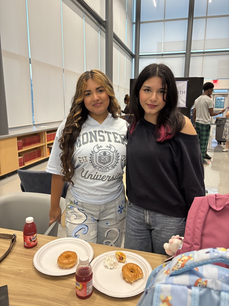 Two women stand behind a table with food. One wears a Monster University shirt. The table holds doughnuts and drinks on white plates.