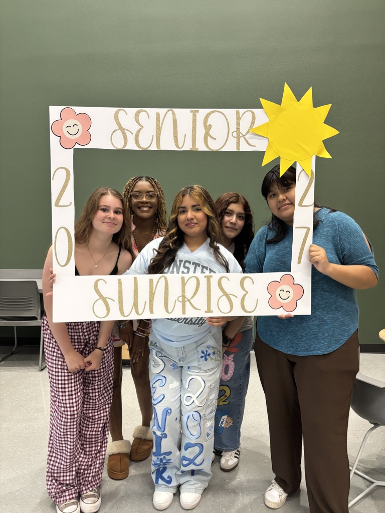 Five women stand together, holding a "Senior 2027 Sunrise" photo frame, smiling. They are in a classroom.
