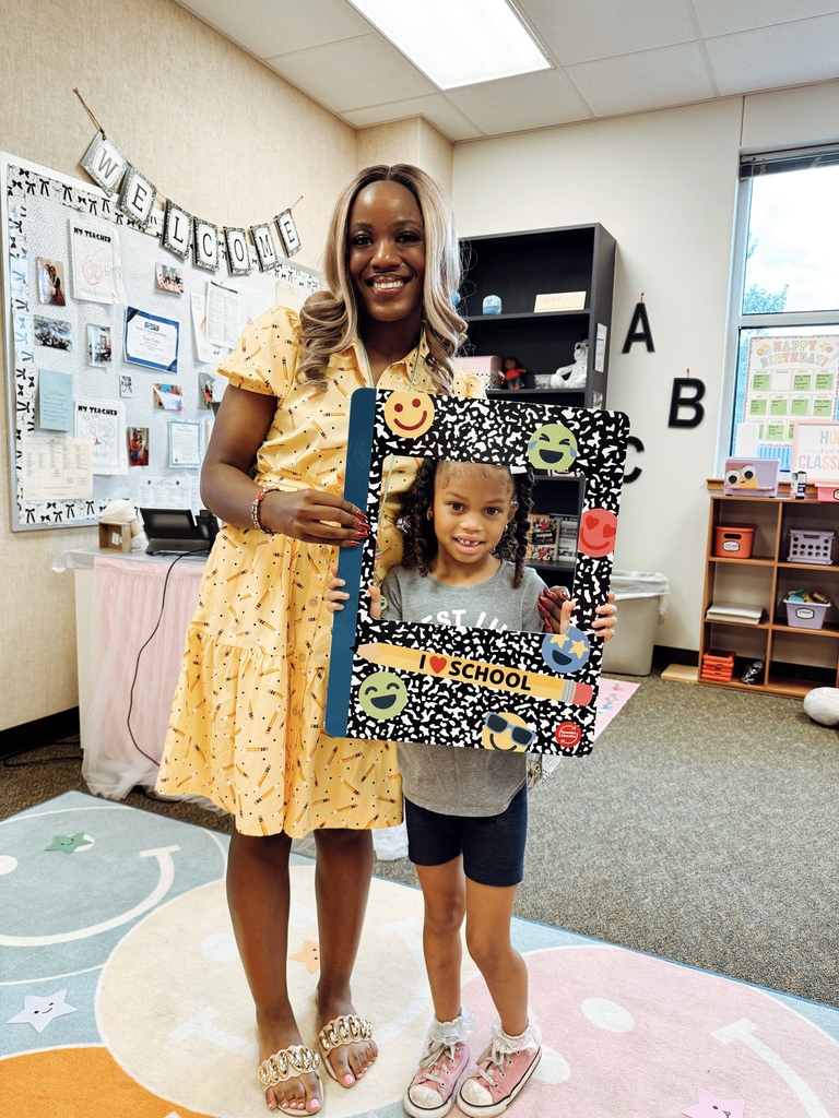 A woman and a child hold a colorful frame with a chalkboard design. They stand in a classroom with bulletin boards and a colorful carpet.