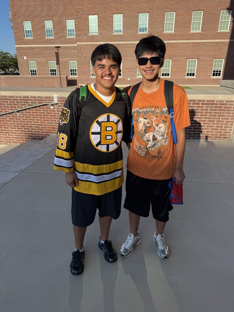 Two young people in casual attire, one in a Boston Bruins jersey, stand outside next to a brick building.