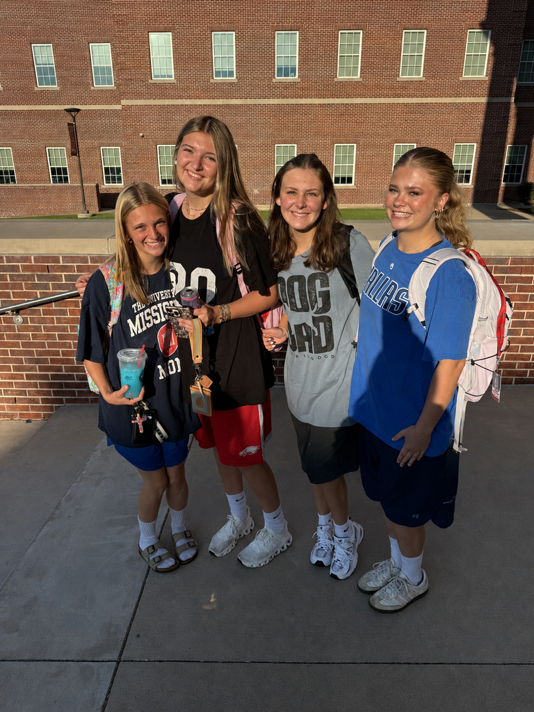 Four young women in casual clothing stand together on concrete with brick building in background.