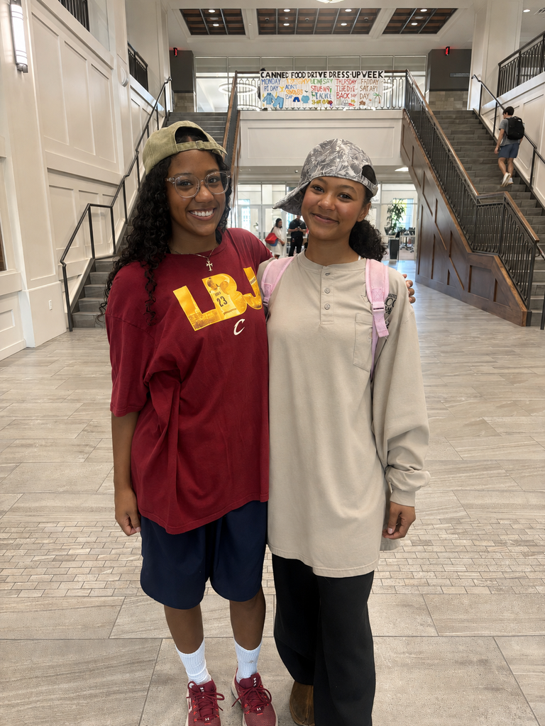Two individuals pose together in a hallway. One wears a maroon shirt and red shoes, the other in a cream shirt and brown boots.