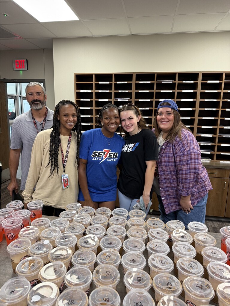 Five people stand next to a table covered with plastic cups. One adult has a badge.