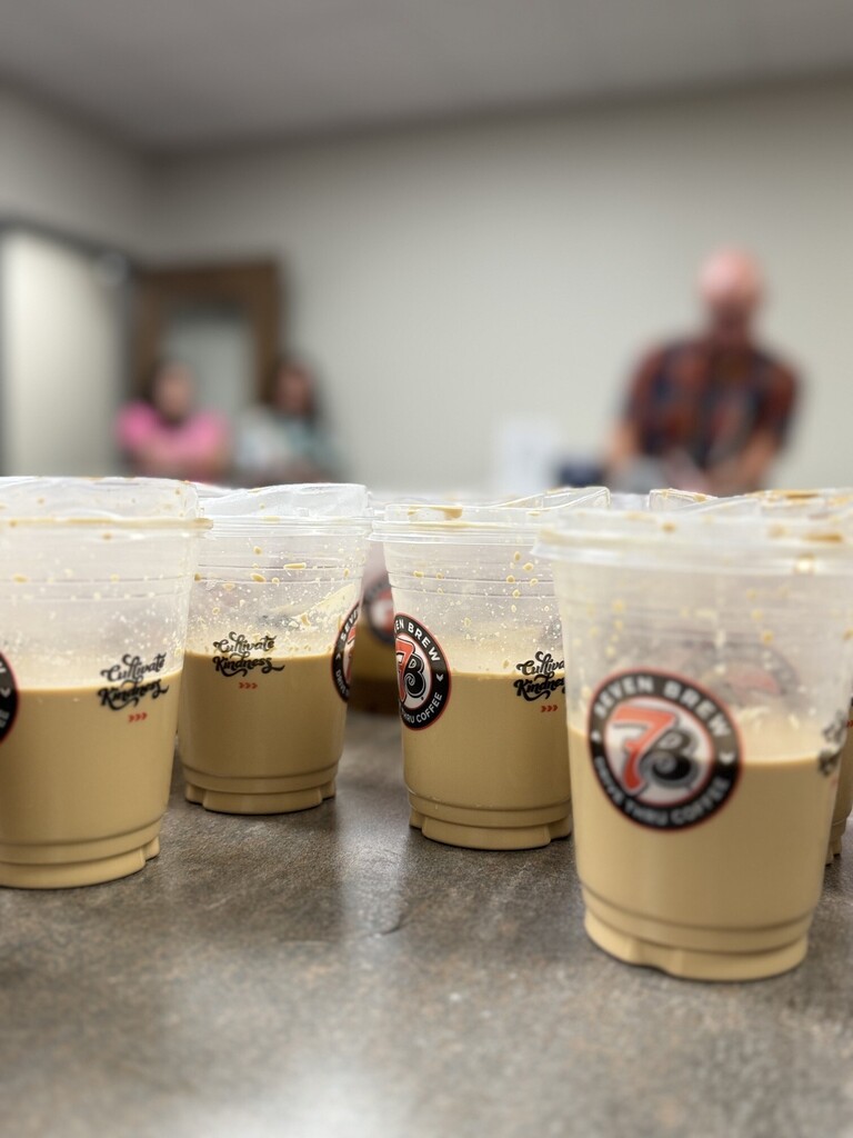 A row of plastic cups with coffee or tea sits on a table. People are in the background.