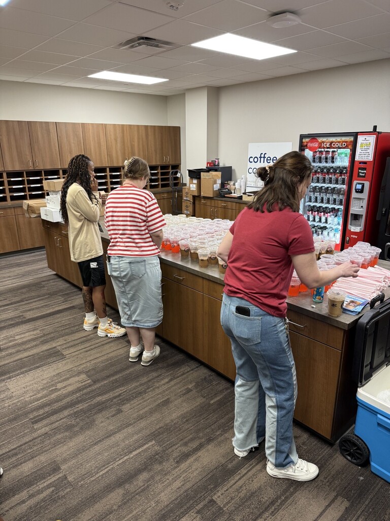 Three individuals gather around a counter in a room with wooden cabinets. They stand before a vending machine and a long table with multiple cups.