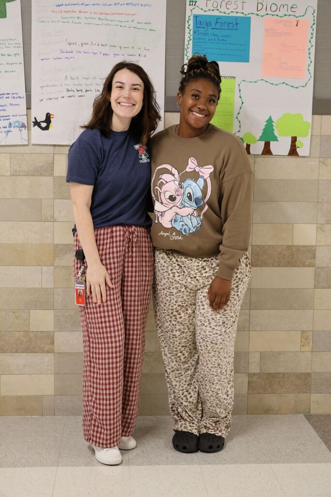 Two women standing close to each other in front of a wall with posters. One wears a blue shirt and red pants; the other wears a brown sweatshirt with a graphic print.